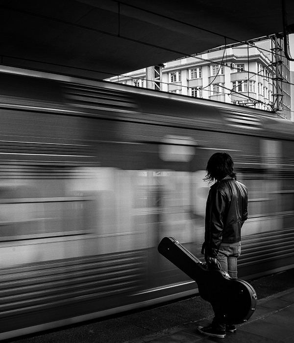 man holding guitar case watching train go by